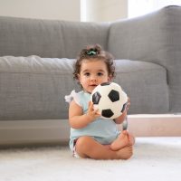 Happy black curly haired baby girl sitting on floor at home Happy black curly haired baby girl in pale blue clothes sitting on floor at home, looking away, playing soccer ball. Front view. Kid at home and childhood concept