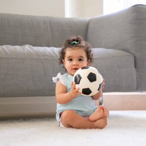 Happy black curly haired baby girl sitting on floor at home Happy black curly haired baby girl in pale blue clothes sitting on floor at home, looking away, playing soccer ball. Front view. Kid at home and childhood concept